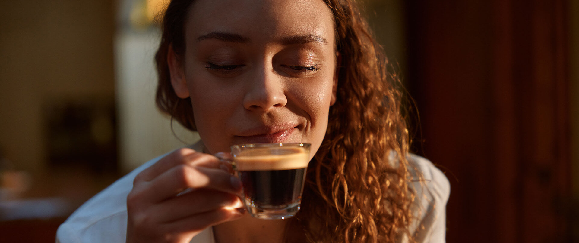 Eine Frau, die einen Espresso in einer Glastasse zum Mund führt und lächelt