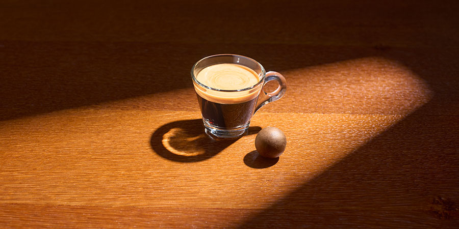 Ein Kaffee in einer Glastasse steht auf einem Tisch, daneben liegt ein Coffee Ball