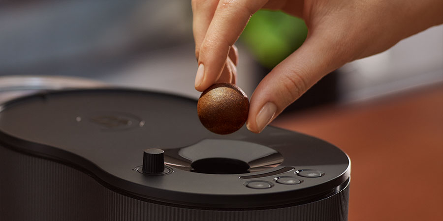 Eine Hand führt einen Coffee Ball zur Öffnung einer Kaffeemaschine